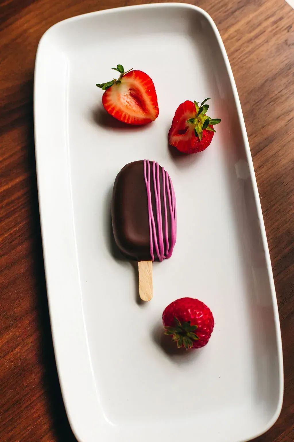 A chocolate cakesicle with pink drizzle on a rectangular white platter, styled with whole and halved strawberries.