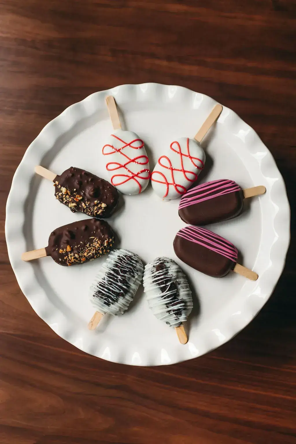 Eight cakesicles arranged in a circle on a round white plate, pairing chocolate-nut, cookies-and-cream, white-with-red-drizzle, and chocolate-with-pink-drizzle designs.