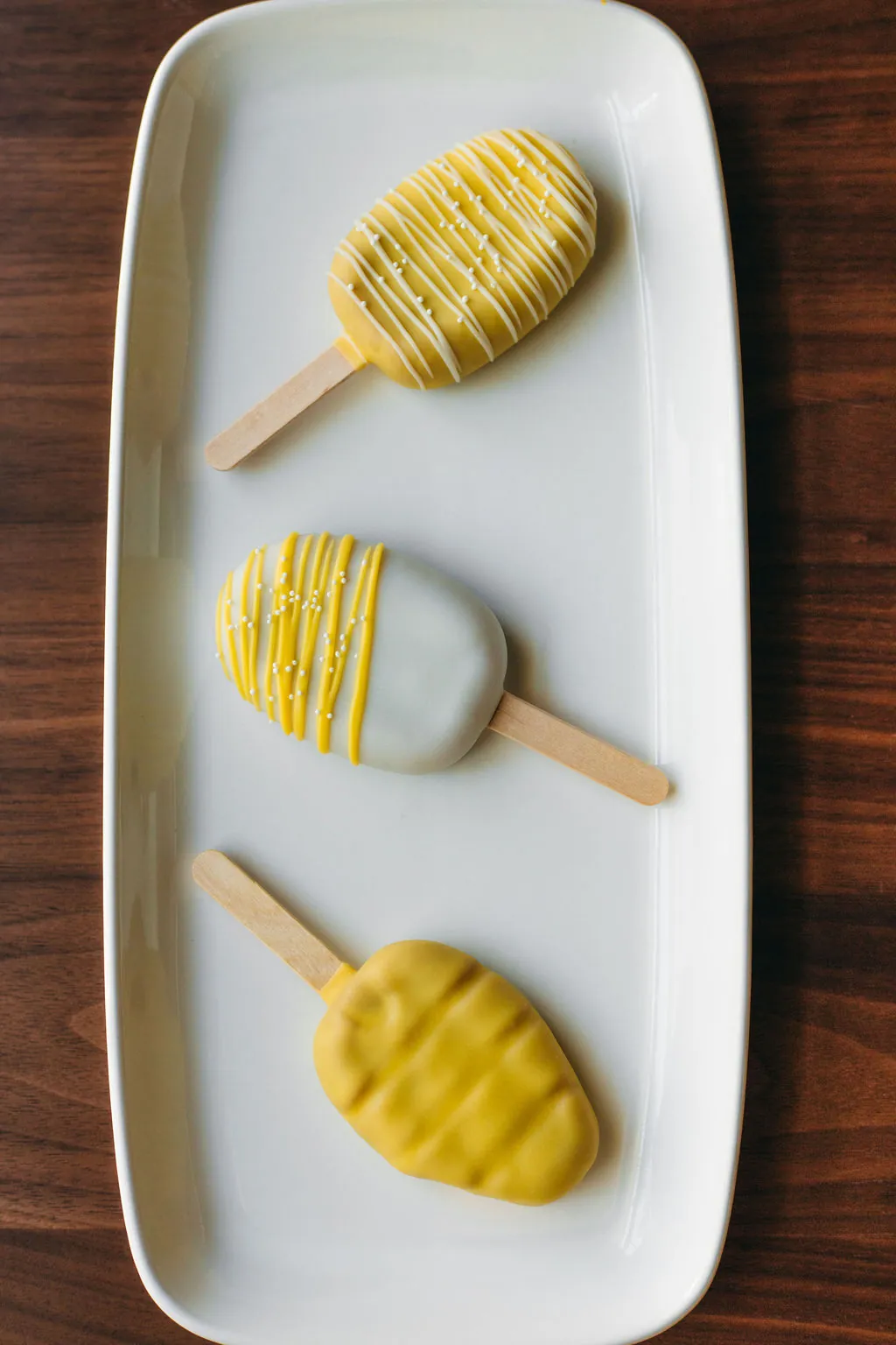 Three yellow and white cakesicles on a rectangular white platter: yellow with white drizzle, white with yellow drizzle, and a honeycomb-textured cakesicle.