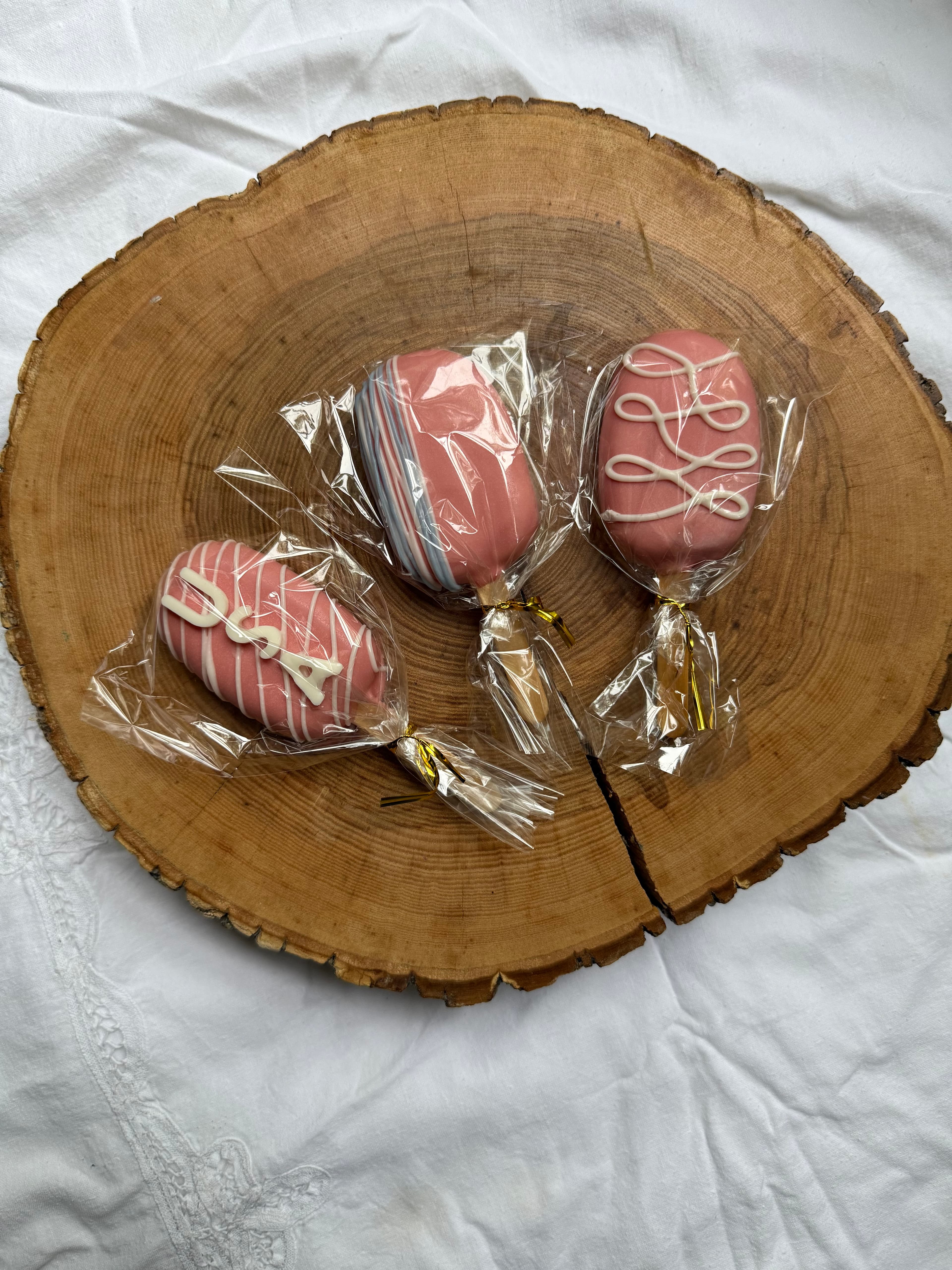 Three blush pink cakesicles with white piped script and stripe designs, individually wrapped and resting on a wood slice.