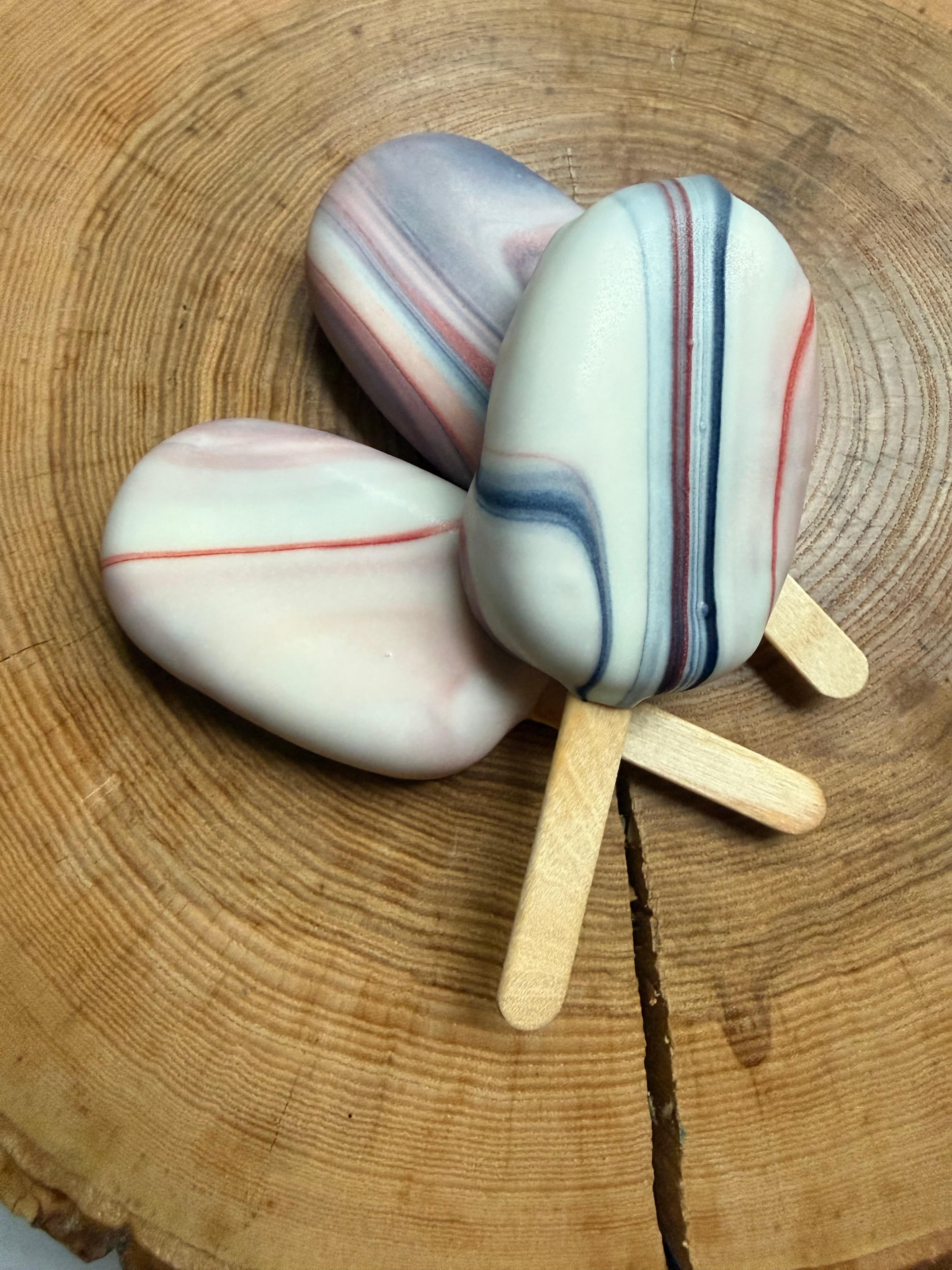 Three cakesicles with a red, white, and blue marbled design, stacked together on a wood slice.