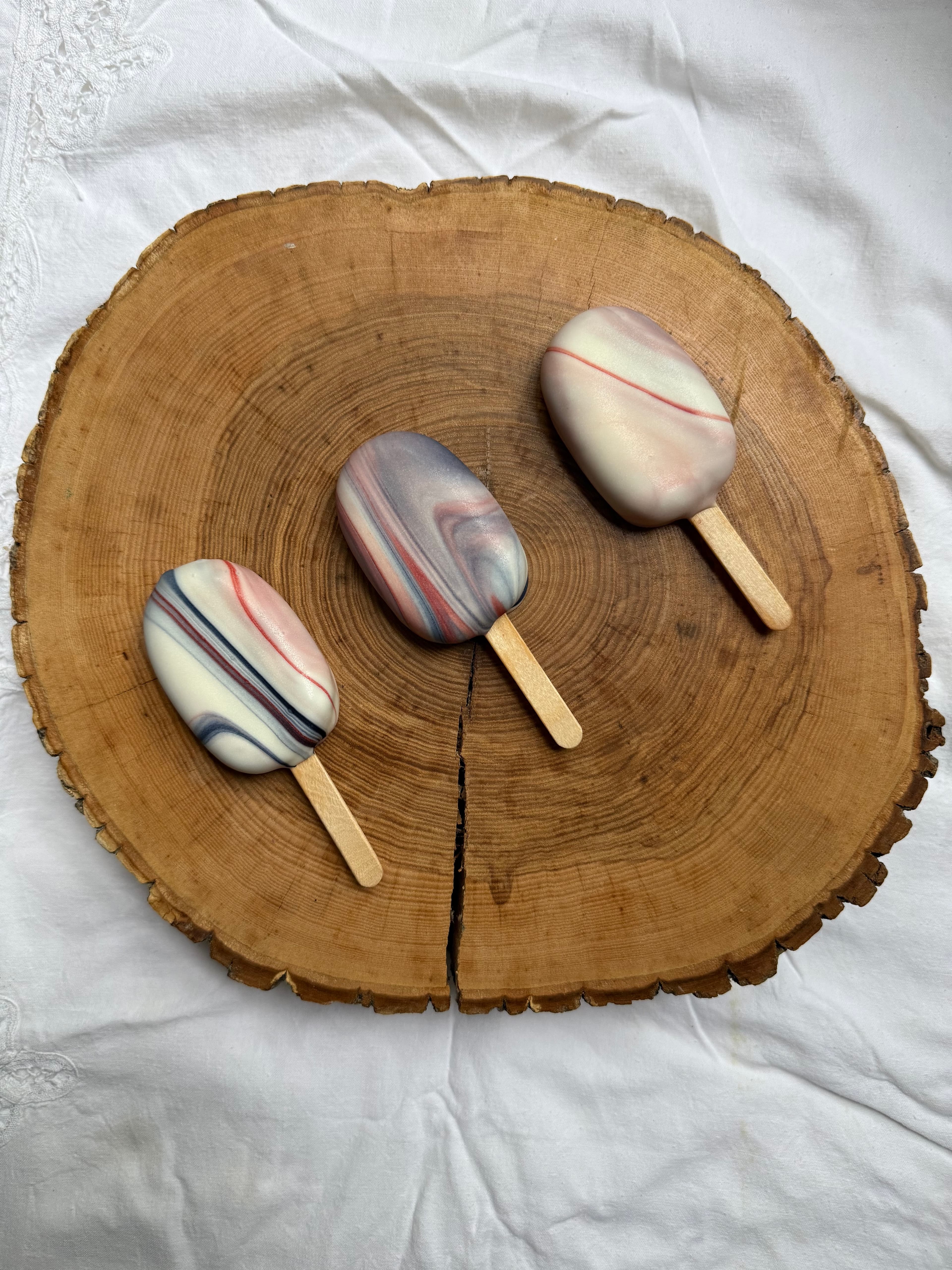 Three cakesicles with a red, white, and blue marbled design, spaced out on a wood slice.