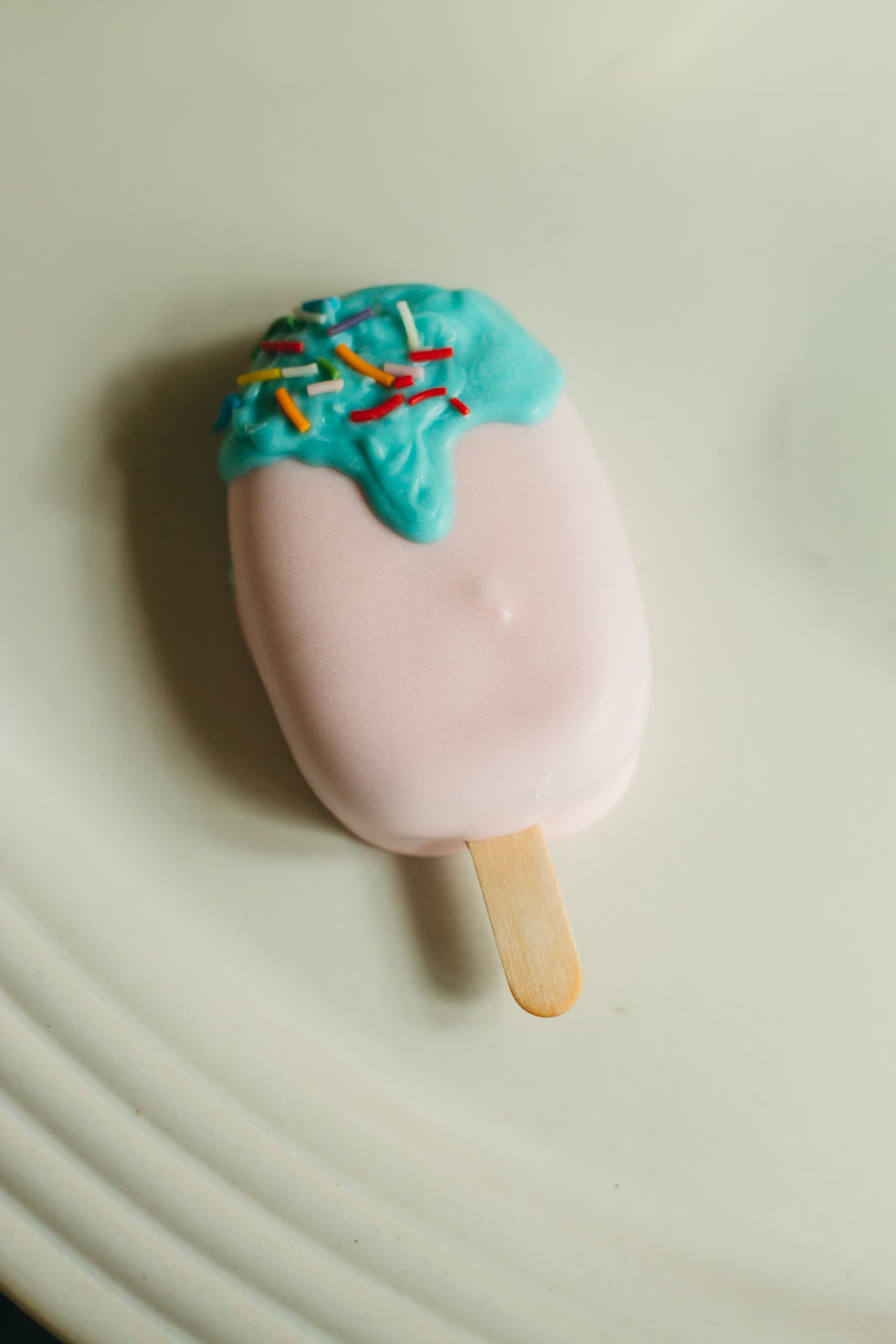 Close-up of a pink cakesicle with a teal drip and rainbow sprinkles on top.