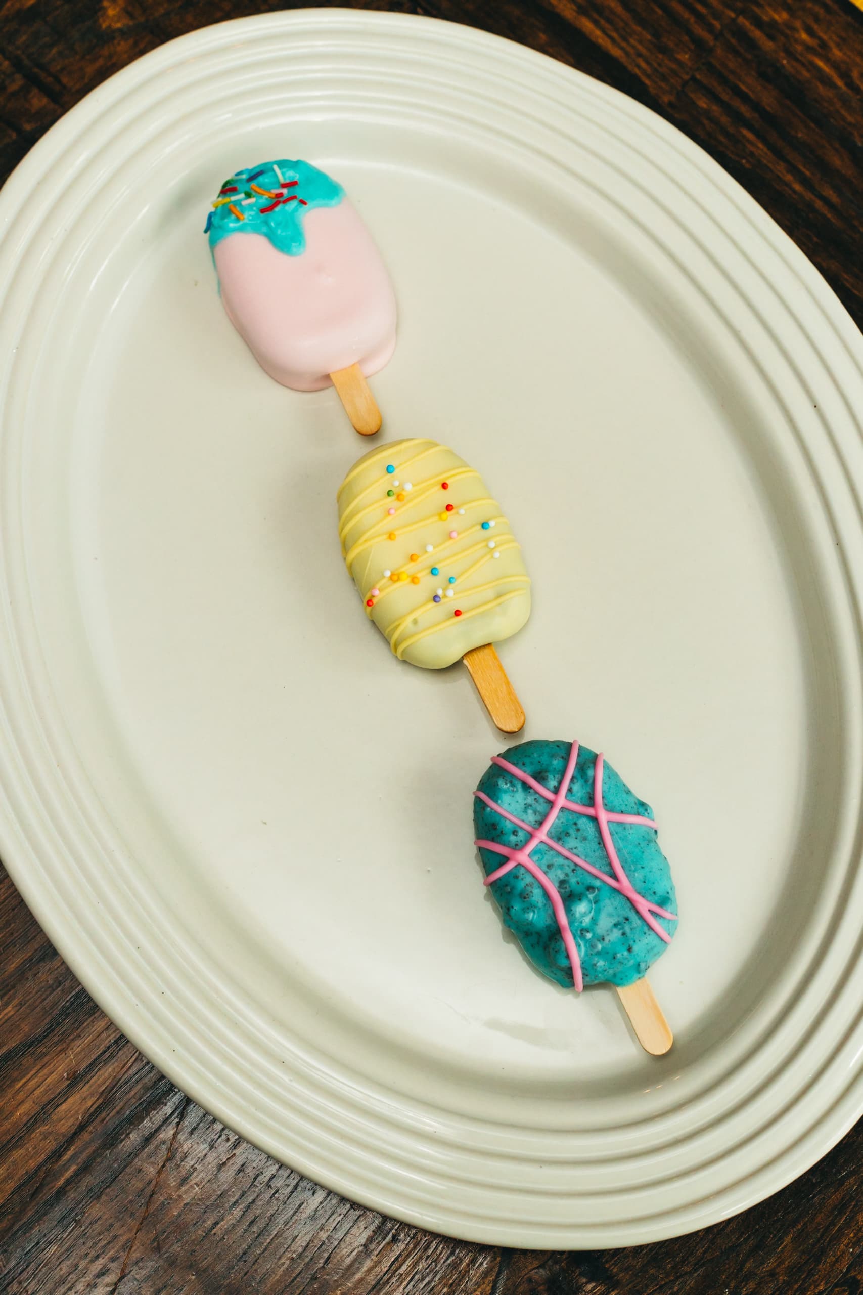 The same pink, yellow, and turquoise cakesicle trio arranged in a row on a wood table.