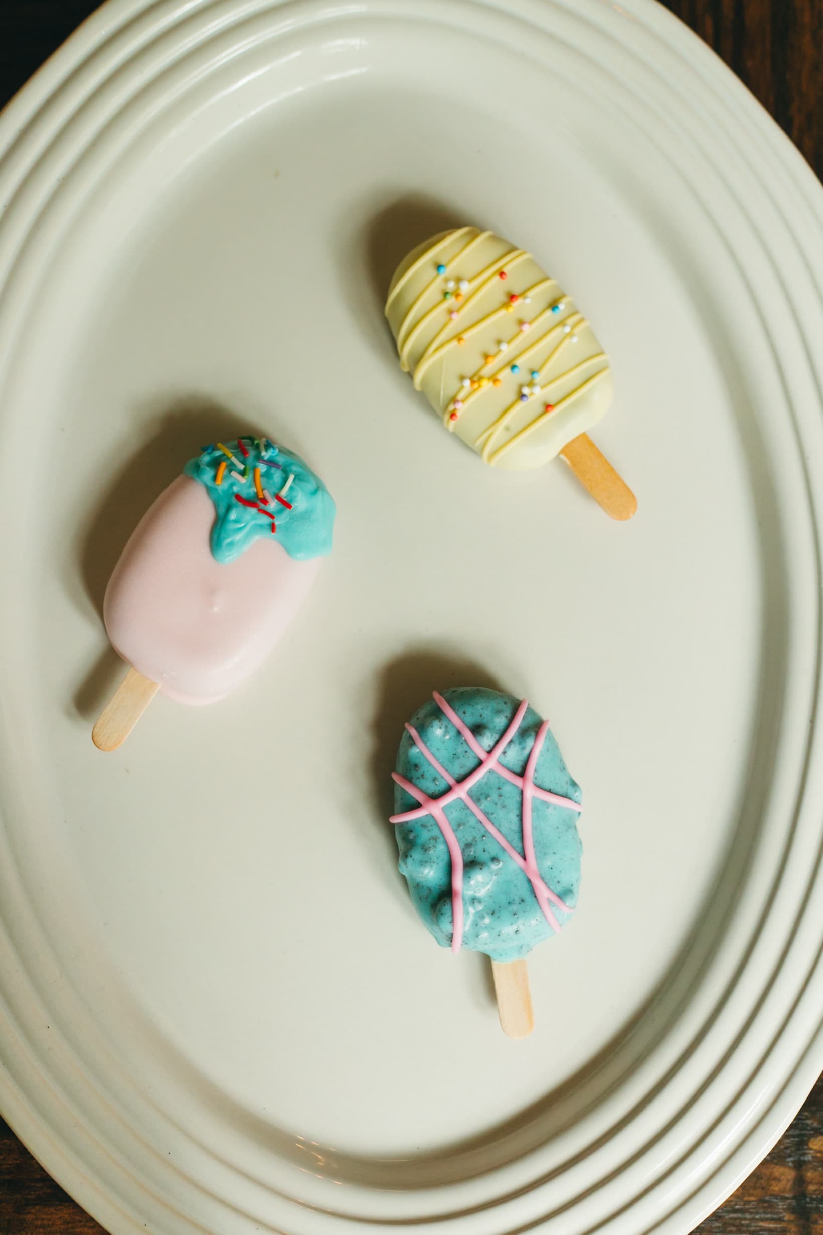 The pink, yellow, and turquoise cakesicle trio arranged in a triangle on a white plate.