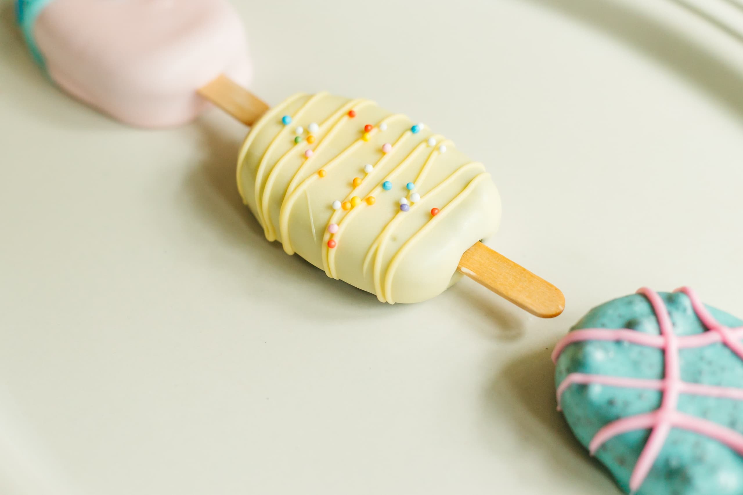 Angled close-up of the pale yellow cakesicle with yellow drizzle and sprinkles, with the pink and turquoise cakesicles blurred in the background.