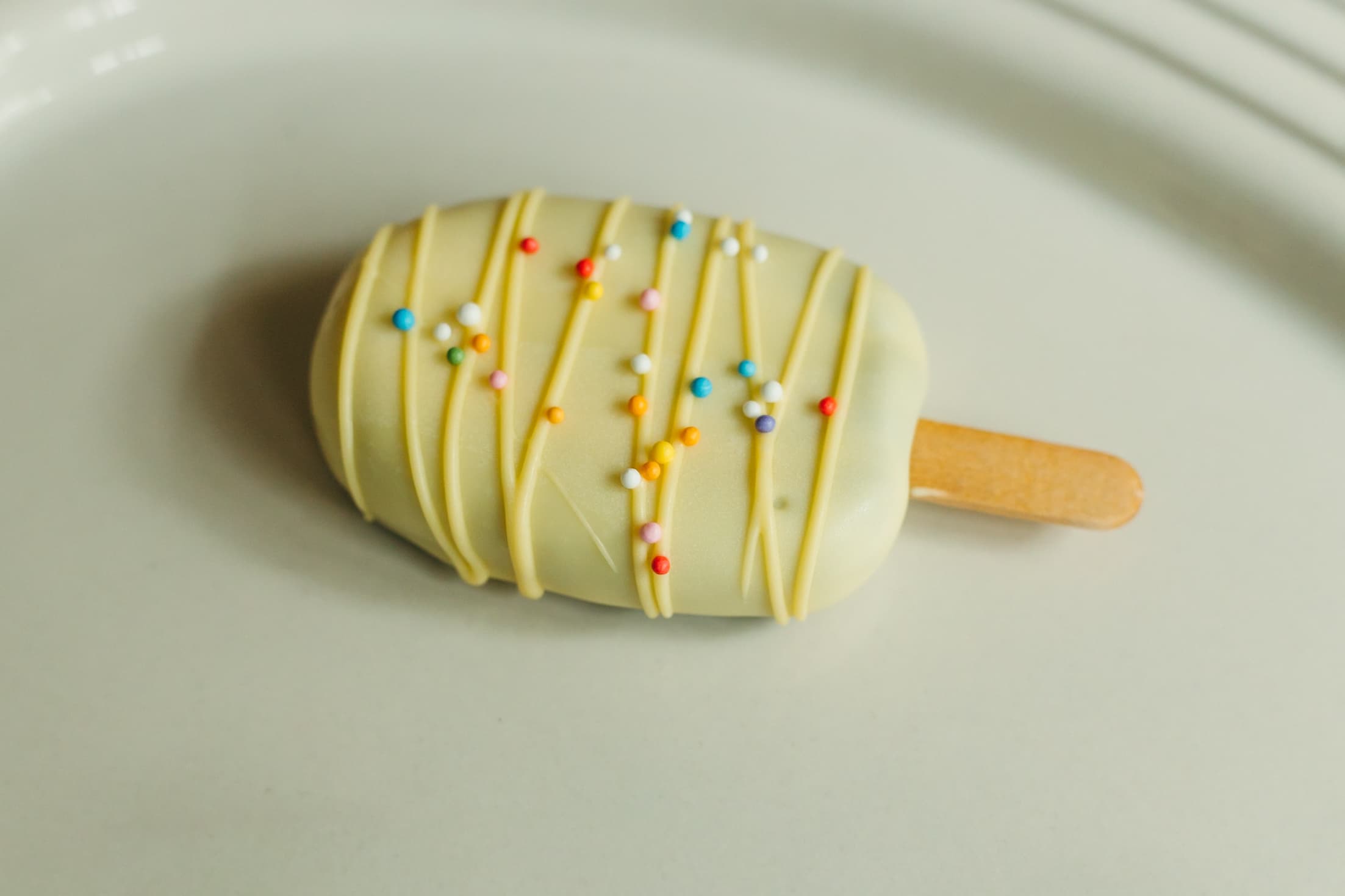 Close-up of a pale yellow cakesicle with yellow drizzle and rainbow sprinkles.