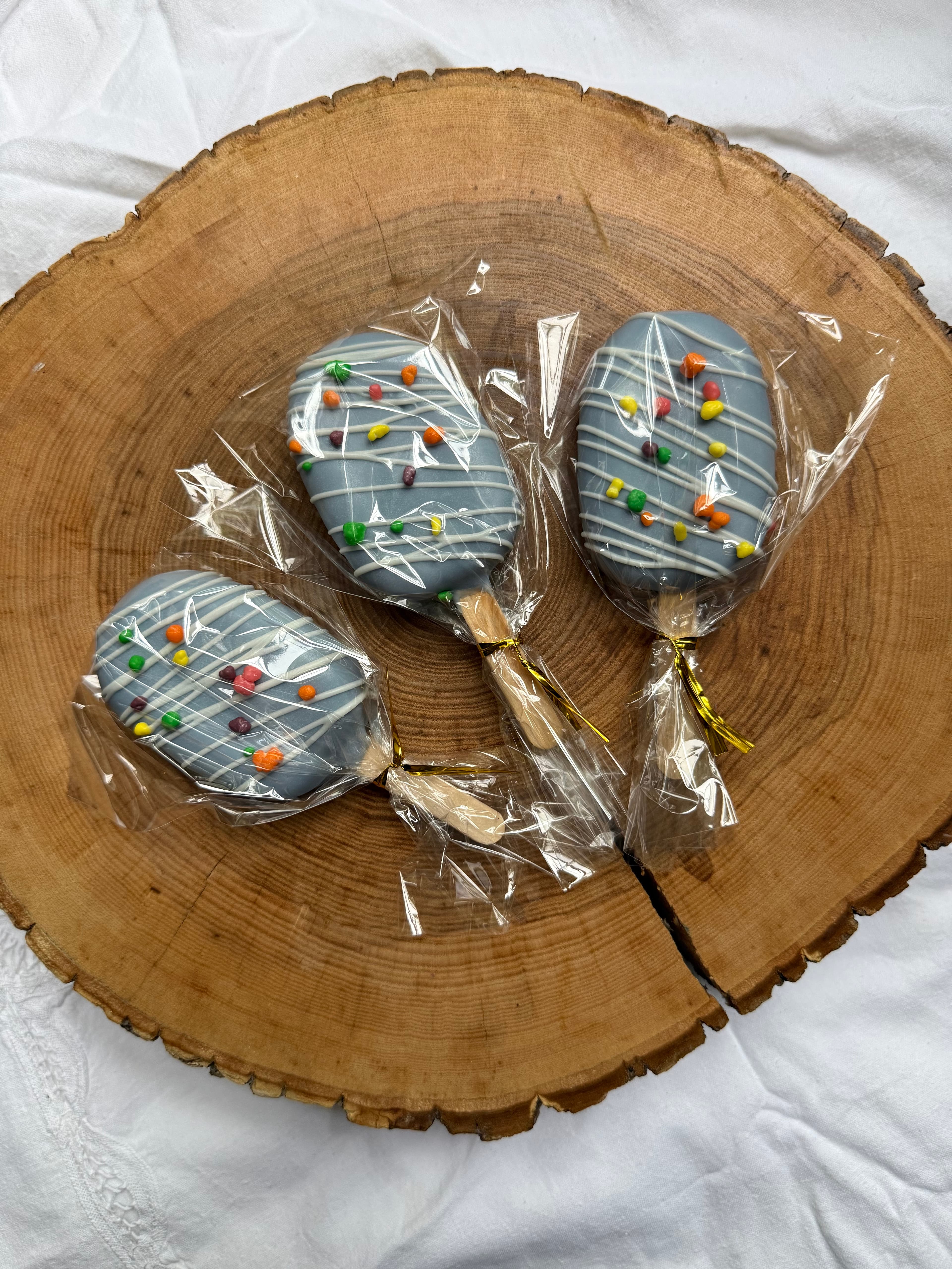 Three gray-blue cakesicles with white drizzle and colorful candy pieces, individually wrapped and resting on a wood slice.